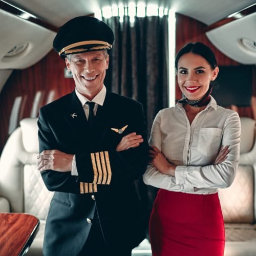 Senior experienced pilot in uniform and beautiful female flight attendant in private jet. Standing in aisle together and meeting passengers